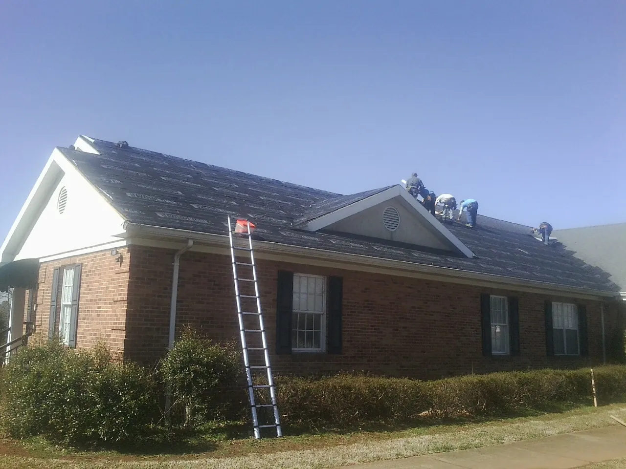 Crew tearing shingles off a brick ranch roof with a ladder set — Emergency Roof Repair work by our crew in Sayville