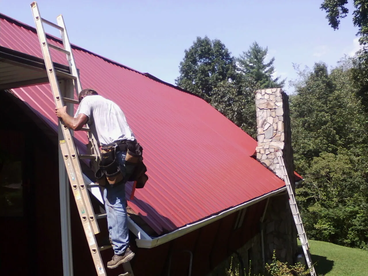 Roofer on a ladder beside a red metal roof and stone chimney — Soffit & Fascia Repair by Roofing Brothers in Sayville